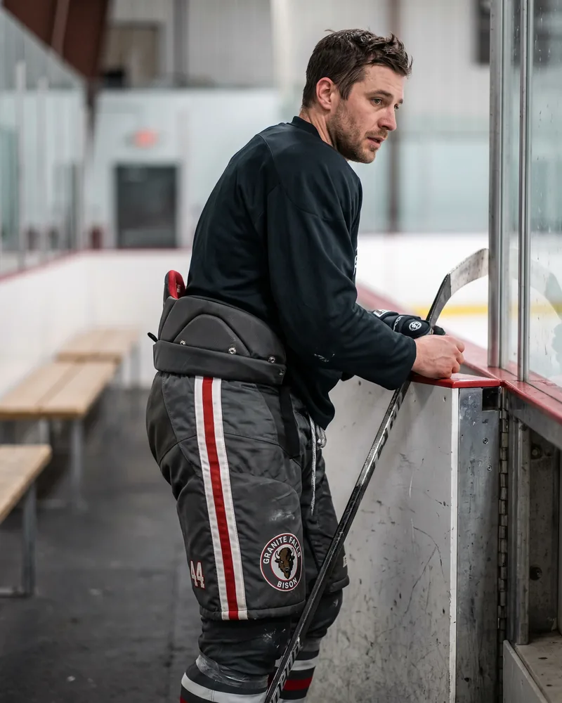 Adult league player at the bench door in charcoal senior hockey pants with a bison crest and number 44