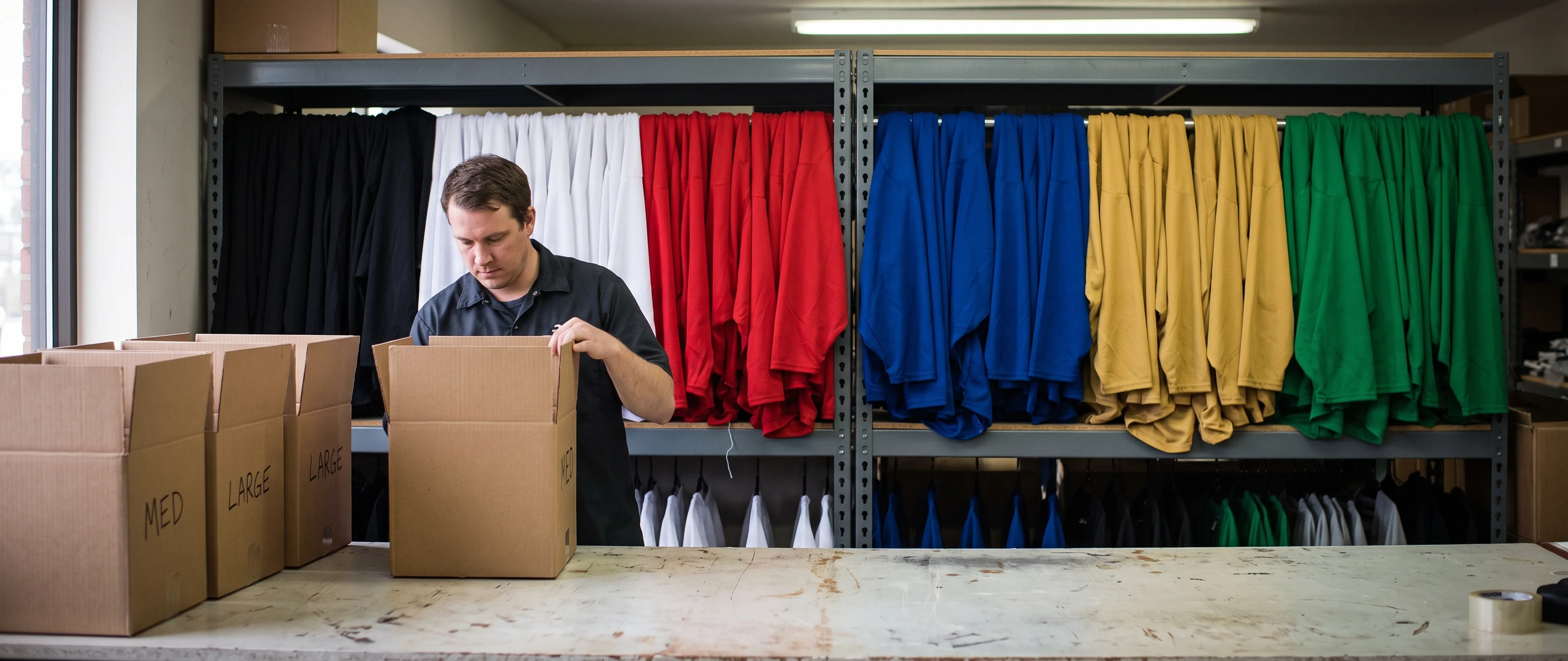 A restock shipment checked into a stockroom beside a rack color-sorted into royal blue, red, black, white, forest green, and gold